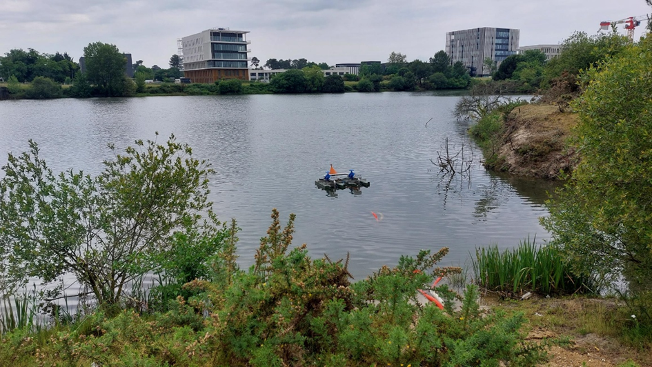Bassin de rétention d’eau pluviale avec dispositif de surveillance flottant, entouré de végétation et de bâtiments modernes en arrière-plan.